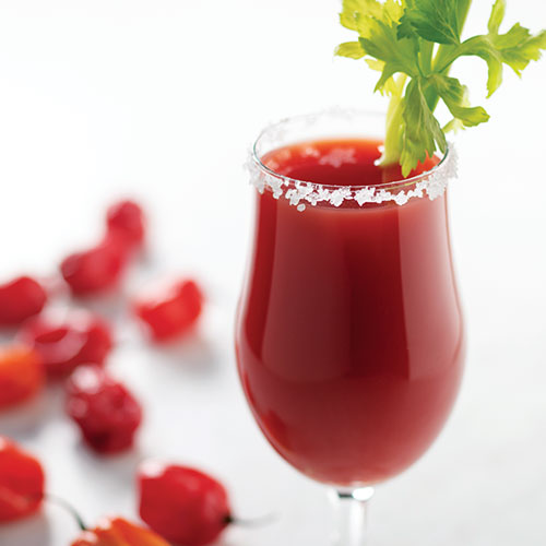 A close up of a Caesar in a poco grande glass garnished with celery and salt on the rim of the glass. Hot peppers on the table in the background.