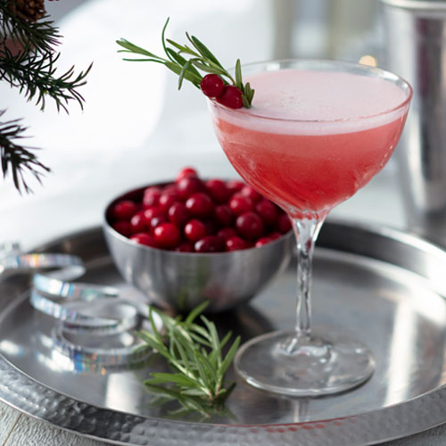 A mixed drink in a coupe glass, garnished with holly and rosemary on a silver platter with a bowl of holly and pine needles.