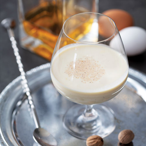 A mixed eggnog drink in a wine glass, topped with spices on a silver platter with a cocktail spoon and walnuts.