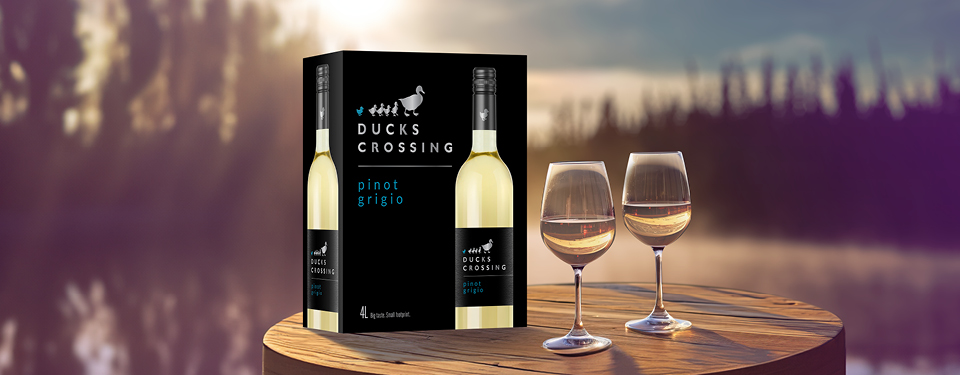 A box of Ducks Crossing Pinot Grigio wine on a table with two wine glasses in front of a lake.